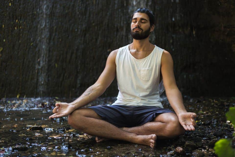 Mann meditiert im Lotossitz an einem Bachwasserfall